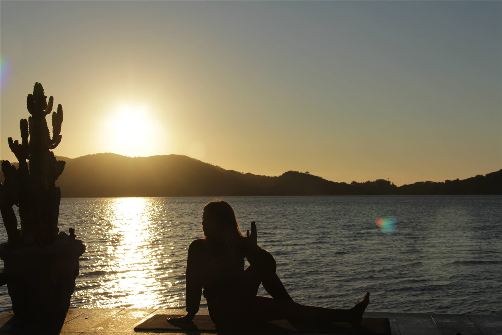 Pessoa meditando em silhueta no deck ao pôr do sol, à beira da lagoa